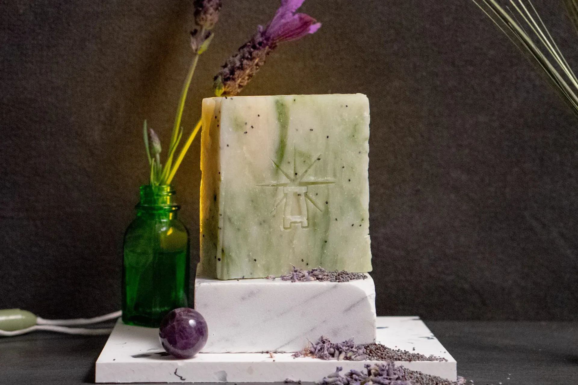 Styled scene with Black Altar green herbal soap on a white marble block, lavender sprigs in a small green glass bottle behind it, loose lavender buds and an amethyst sphere arranged in front.