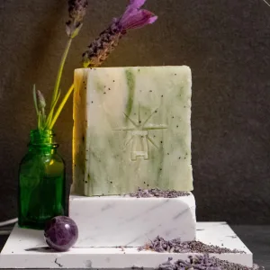 Styled scene with Black Altar green herbal soap on a white marble block, lavender sprigs in a small green glass bottle behind it, loose lavender buds and an amethyst sphere arranged in front.