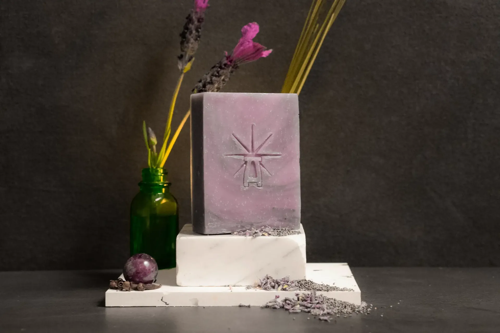 Styled scene featuring Black Altar lavender-hued Mystic Twilight soap on stacked white marble blocks, with dried lavender stems in a green glass bottle behind it and scattered lavender buds and an amethyst sphere in front.