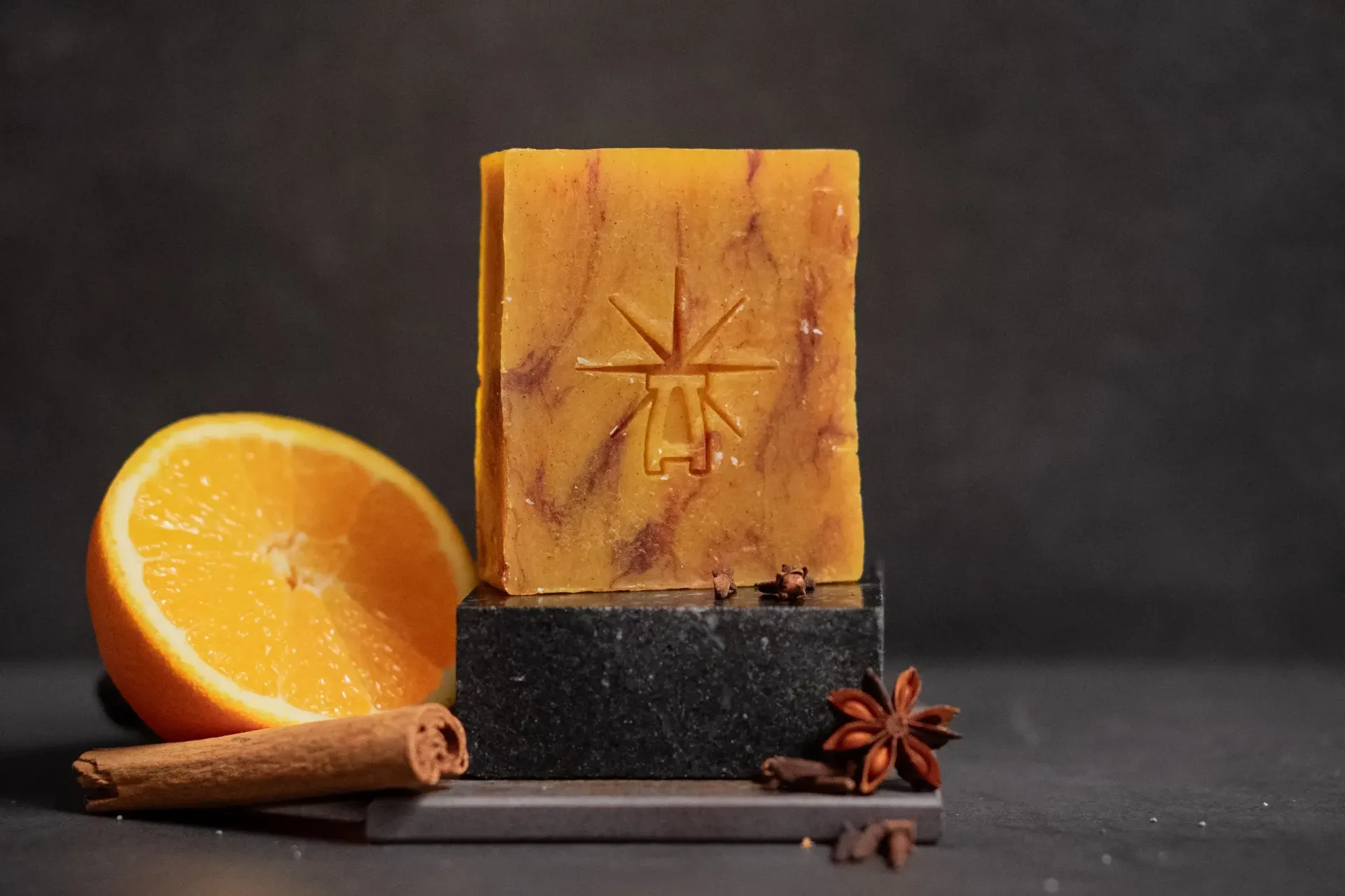 Black Altar Ritual Ember soap—warm amber bar on a dark stone pedestal, framed by a sliced orange, cinnamon stick, star anise, and cloves.