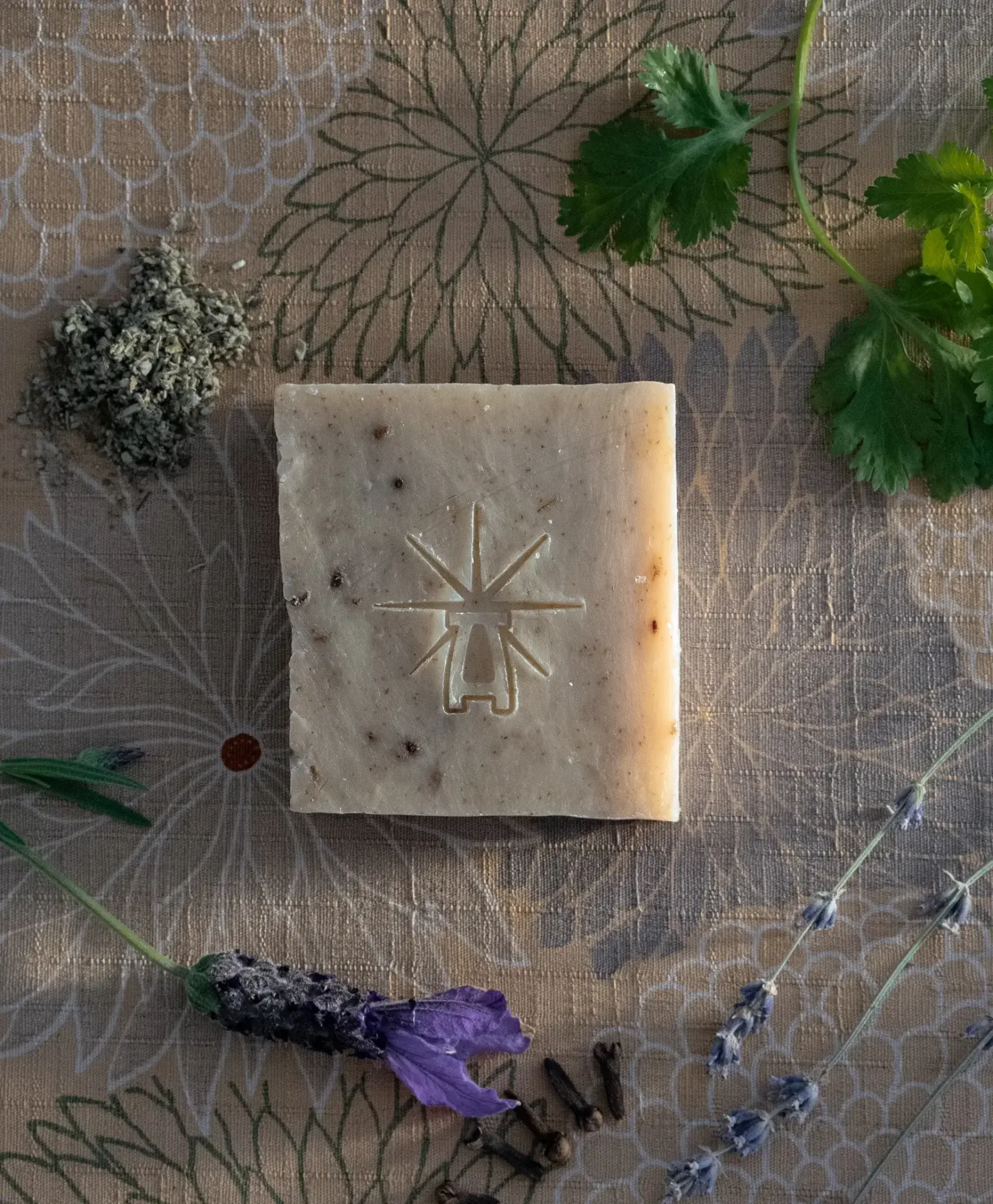 Flat-lay of Black Altar Witch Bloom soap surrounded by dried sage, fresh parsley, lavender flower, and cilantro leaves on floral fabric.
