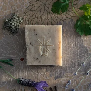 Flat-lay of Black Altar Witch Bloom soap surrounded by dried sage, fresh parsley, lavender flower, and cilantro leaves on floral fabric.