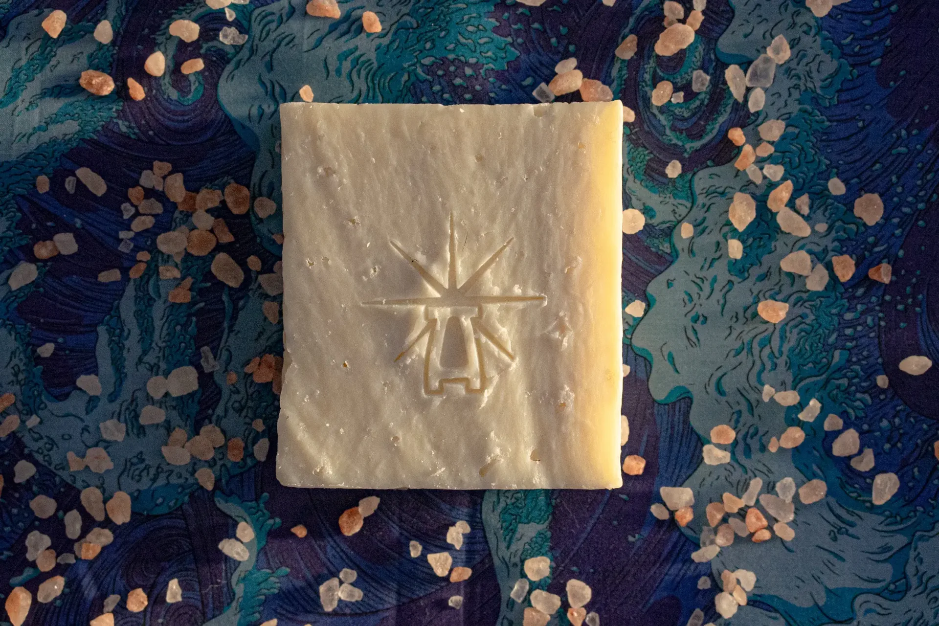 Flat-lay of Black Altar Earth Salt soap on ocean-blue fabric, scattered with coarse pink Himalayan salt crystals.