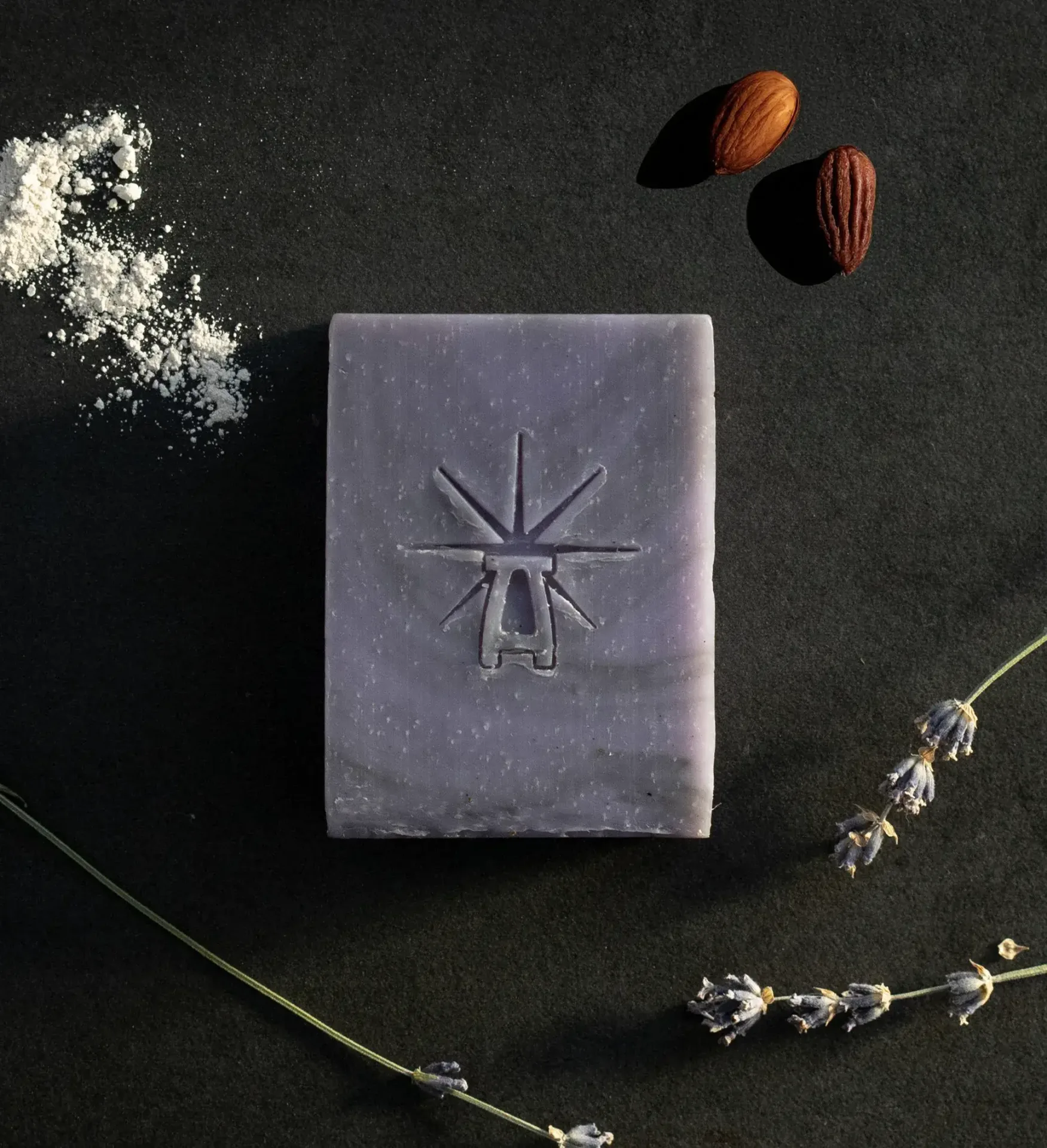 Black Altar Mystic Twilight soap on dark surface, accented by powdered clay, two shea butter nuts, and dried lavender stems.