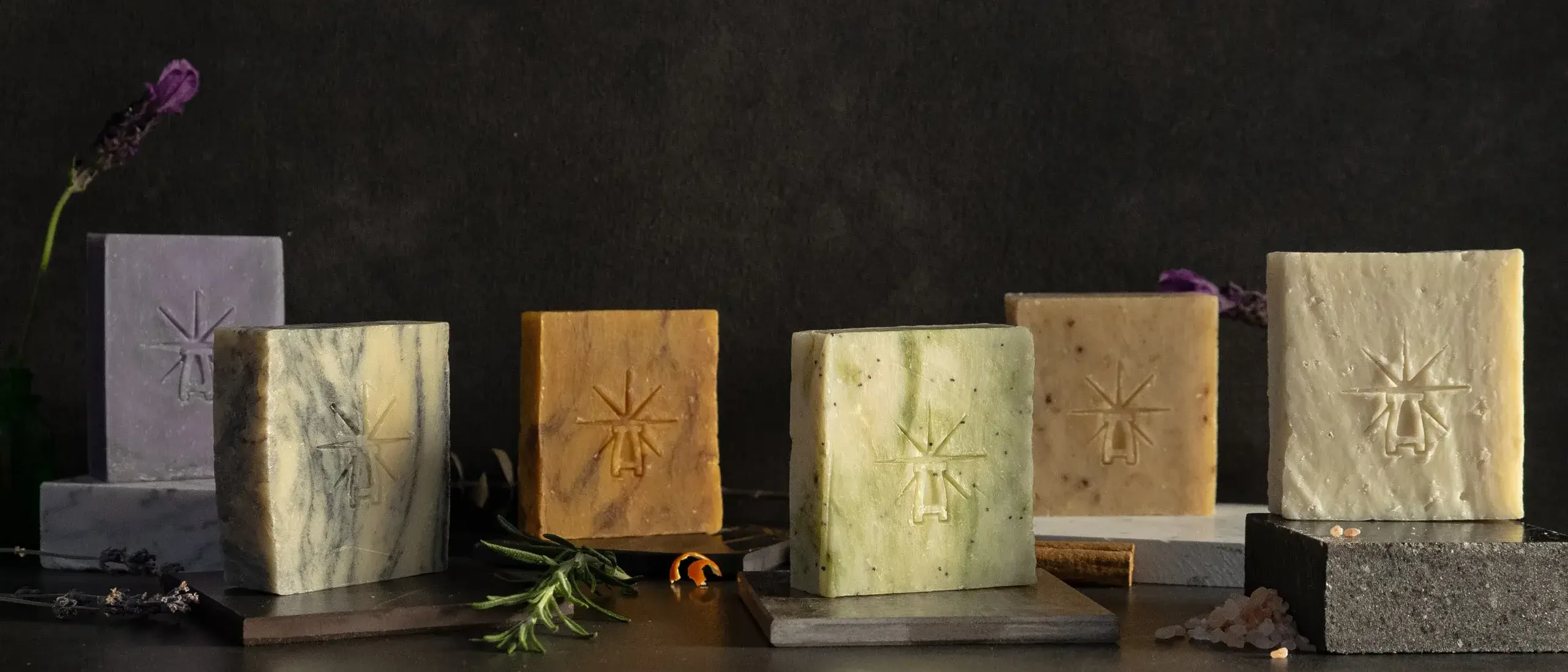 Black Altar Soap Line-up Six Black Altar soap bars in varied colors—lavender, marble, amber, green, beige, and salt white—displayed with herbs and spices against a dark background.