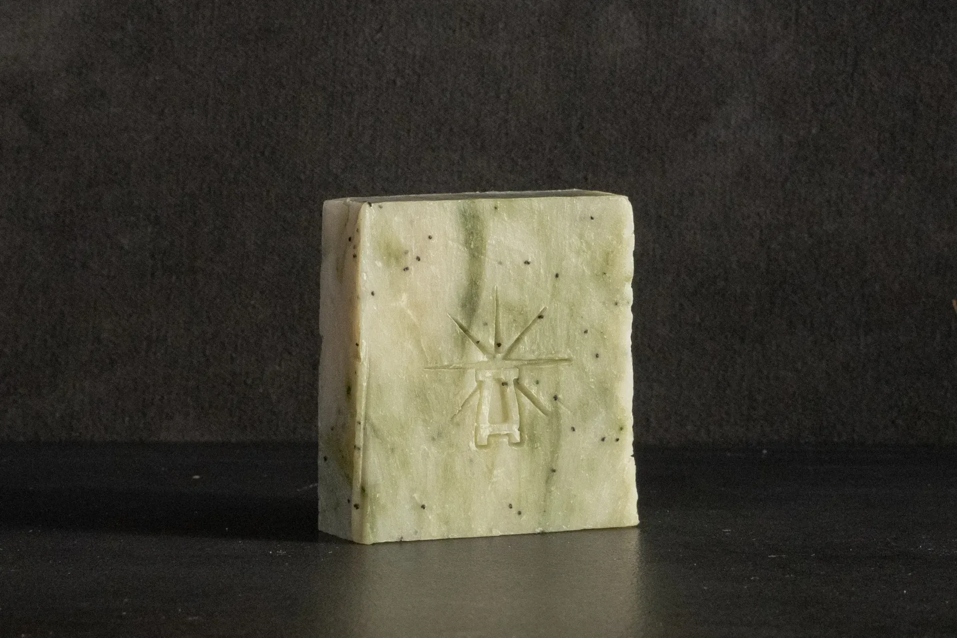 Studio shot of Black Altar Wild Conjuring soap—pale green bar with dark poppy specks and star-altar logo on dark backdrop.