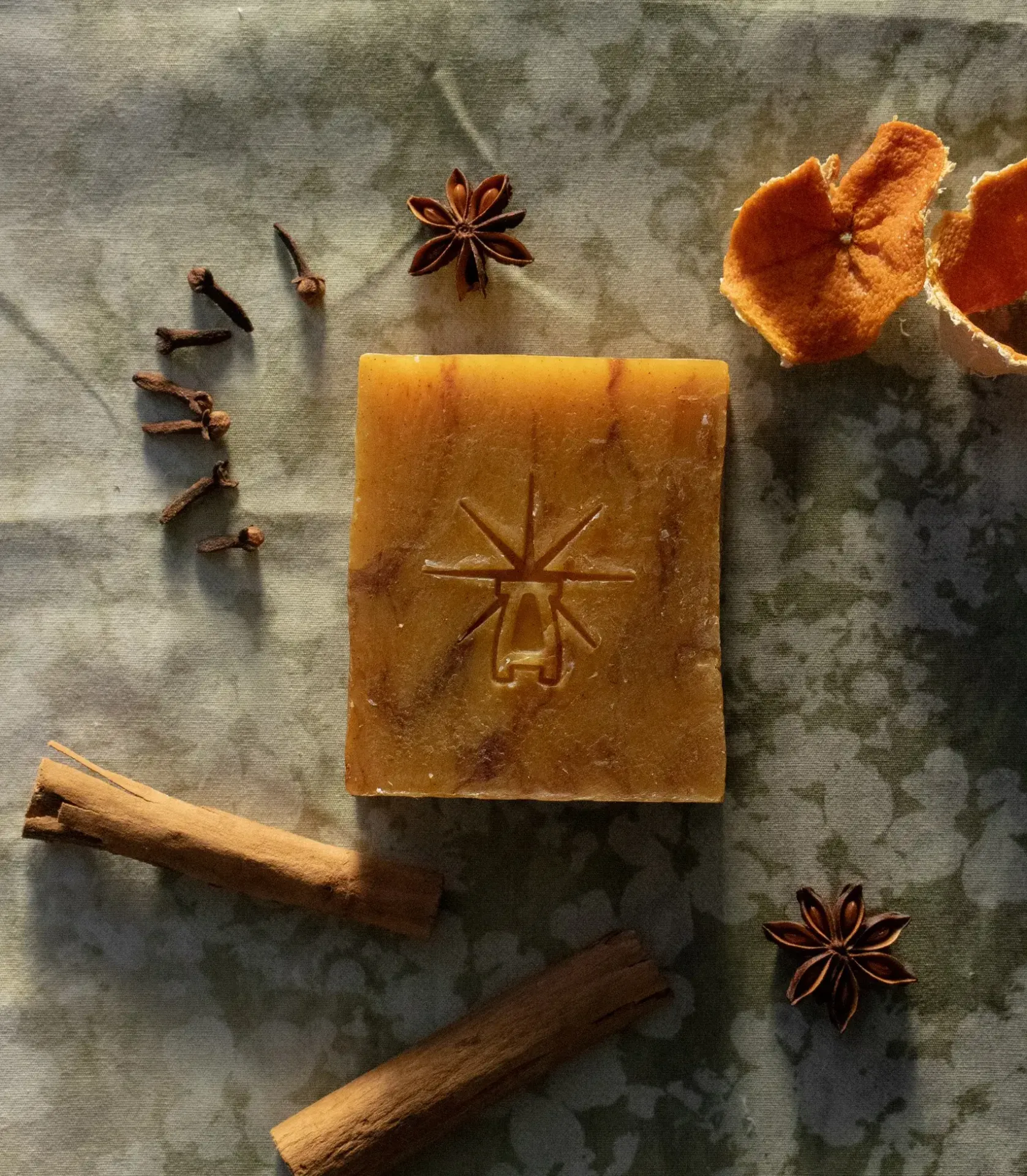 Flat-lay of Black Altar Ritual Ember soap with cloves, star anise, cinnamon sticks, and dried orange peel on mottled green fabric.