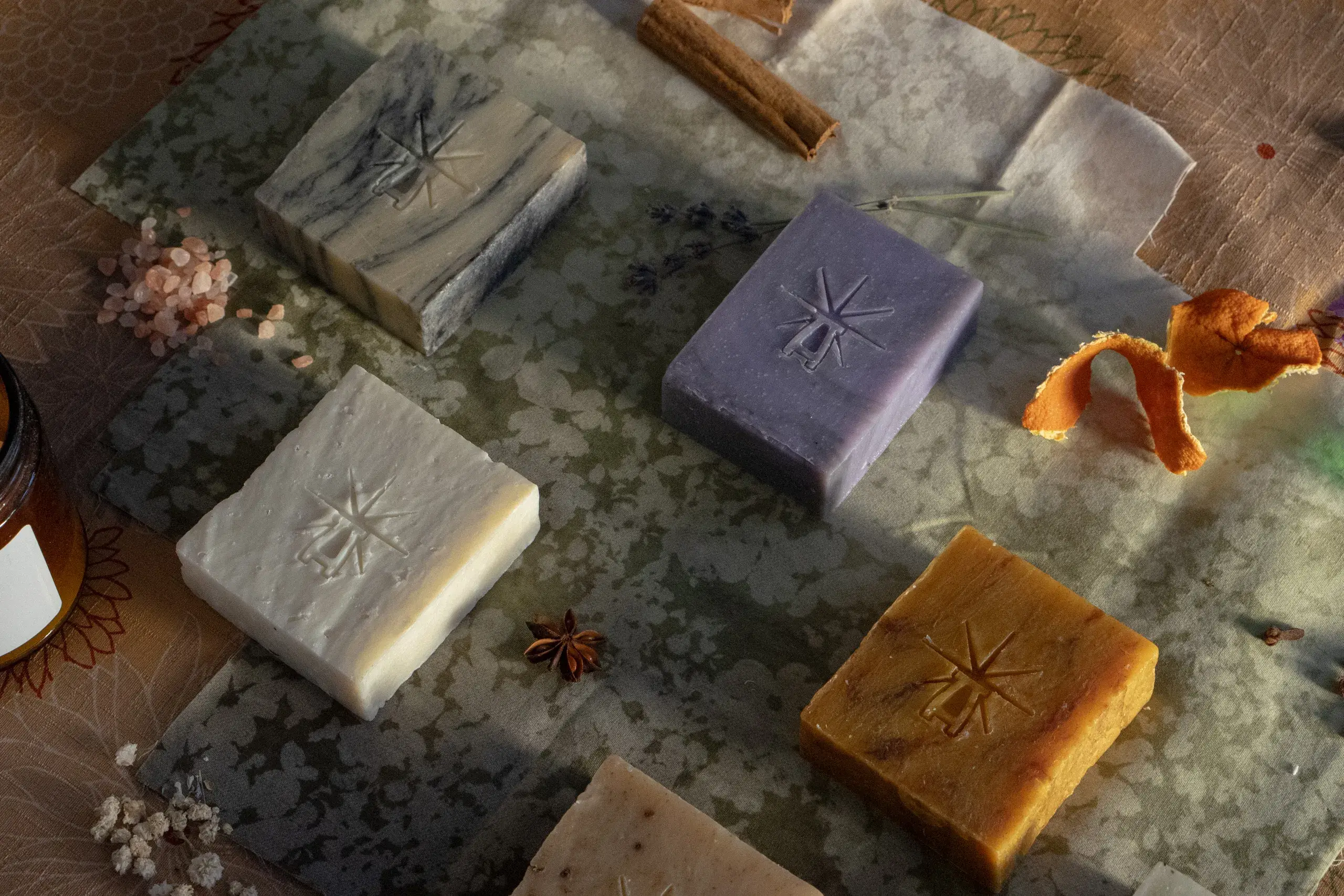 Angled overhead view of five Black Altar soaps—marble, lavender, salt white, amber, and salt crystals—arranged on layered fabric with cinnamon stick, dried lavender, orange peel, and Himalayan salt.