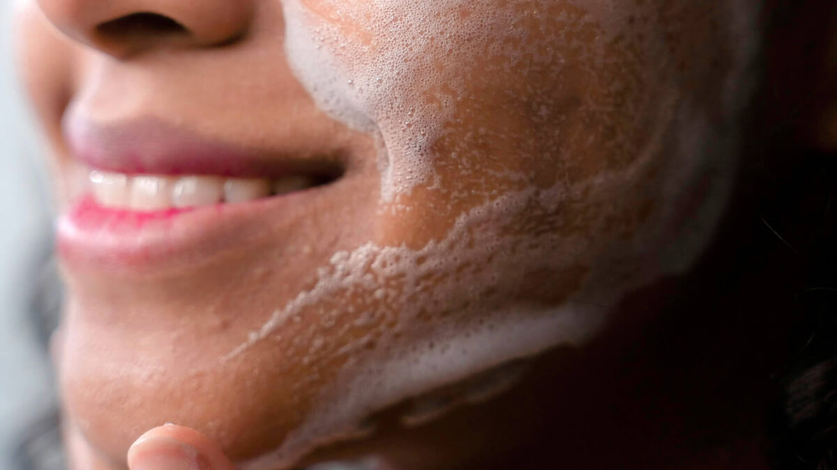 woman with soap suds on her face smiling while rubbing her chin.
