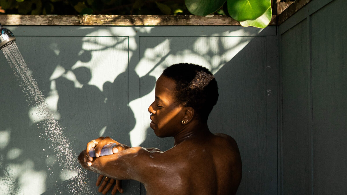 Phillip showering outdoors, sun-dappled, lathering arm with a bar of soap. twilight charcoal—lavender charcoal