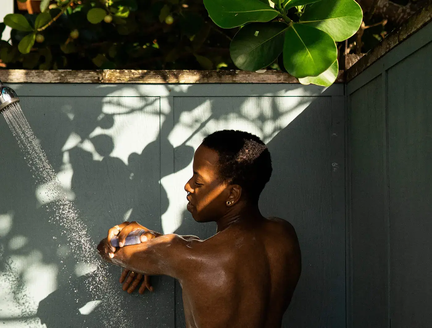Phillip showering outdoors, sun-dappled, lathering arm with a bar of soap. twilight charcoal—lavender charcoal