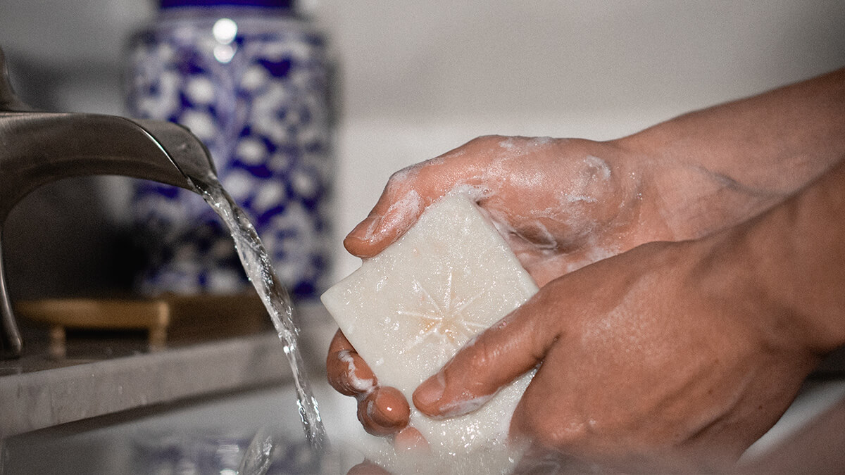 Sierra washes her hands with a salt scrub bar that shows the Black Altar A symbol on it. there's a gold soap dish, a blue floral vase and water is running.