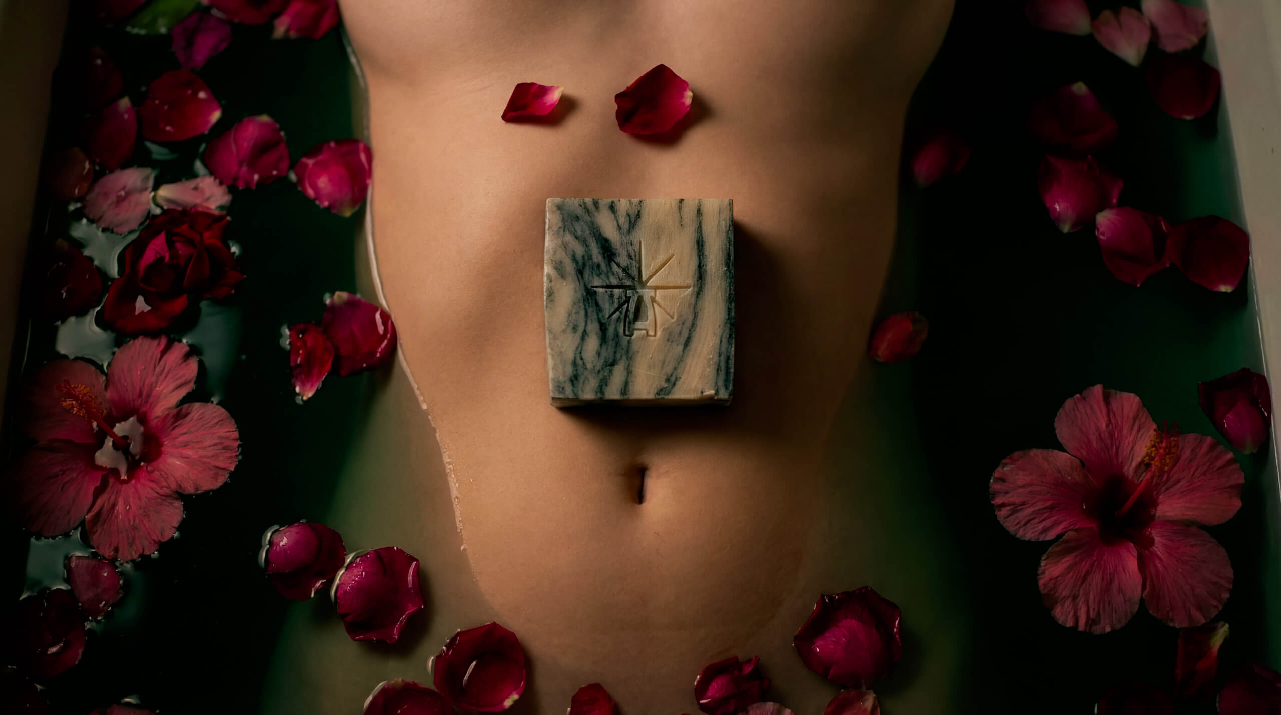A woman lies in a bath filled with water surrounded by flower petals. Theres a bar of Marbled Charcoal sitting on her belly above her belly button.